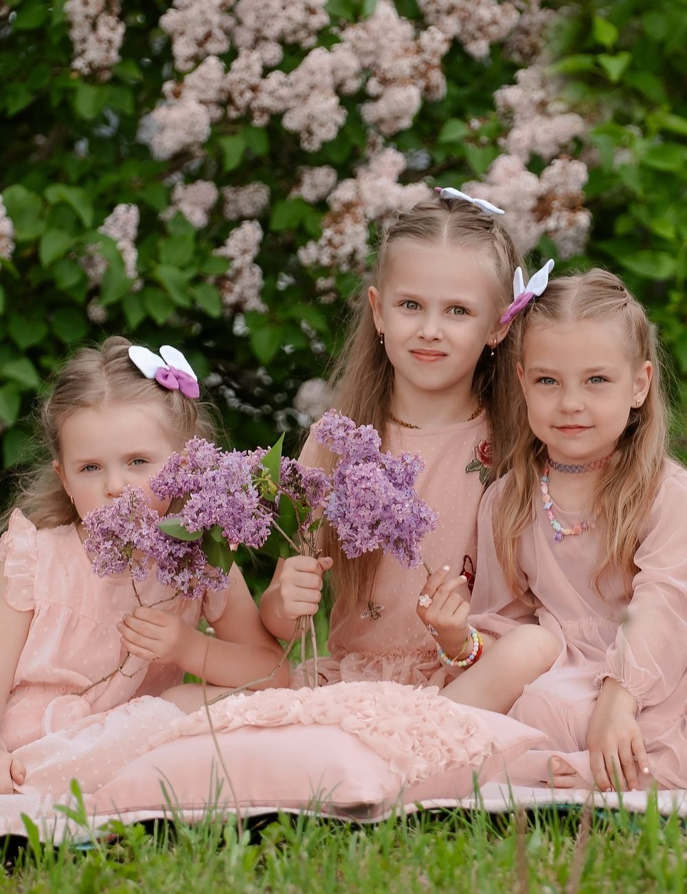 Trys mergaitės rausviomis suknelėmis laiko alyvų šakeles sode – vaikų fotosesija Šiauliuose