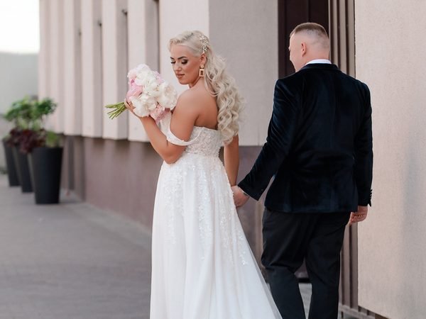 Nuotaka ir jaunikis eina susikibę rankomis, ji žvelgia atgal su puokšte – vestuvių fotosesija Šiauliuose