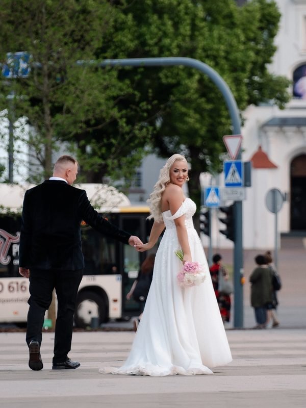 Jaunieji susikibę už rankų eina per miesto gatvę su puokšte – vestuvių fotosesija Šiauliuose