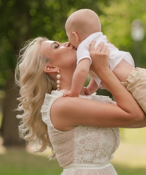 Mama pakėlusi kūdikį bučiuoja jį, aplinkui žalia gamta vasaros dieną – krikštynų fotosesija Šiauliuose