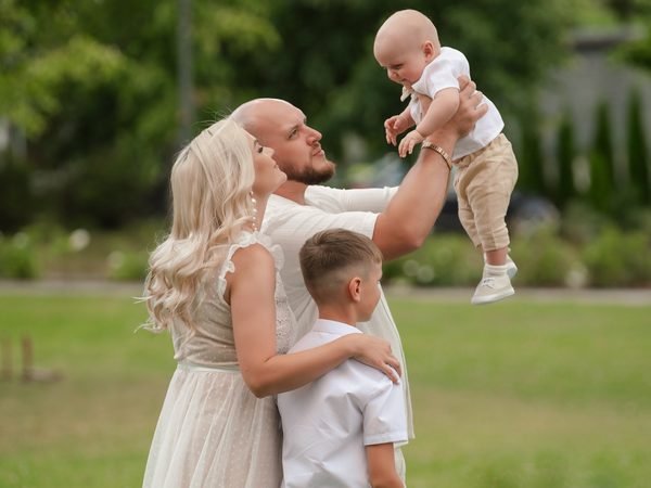 Tėtis pakelia kūdikį aukštyn, šalia mama ir berniukas parke – krikštynų fotosesija Šiauliuose