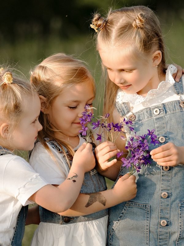 Trys mergaitės pievoje kvepia violetinėmis laukų gėlėmis – vaikų fotosesija Šiauliuose