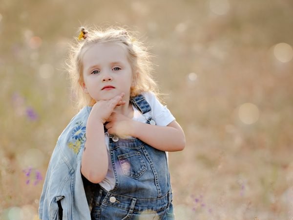 Mergaitė džinsiniu kombinezonu pozuoja pievoje saulėlydžio šviesoje – vaikų fotosesija Šiauliuose