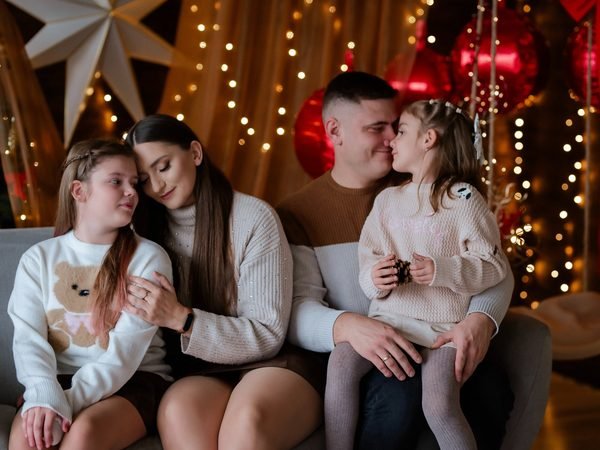 Šeima su dviem dukromis apsikabinusi sėdi ant sofos kalėdinėje aplinkoje – kalėdinė fotosesija Šiauliuose