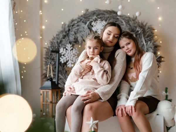 Mama apkabinusi dvi dukras sėdi tarp kalėdinių papuošimų – kalėdinė fotosesija Šiauliuose