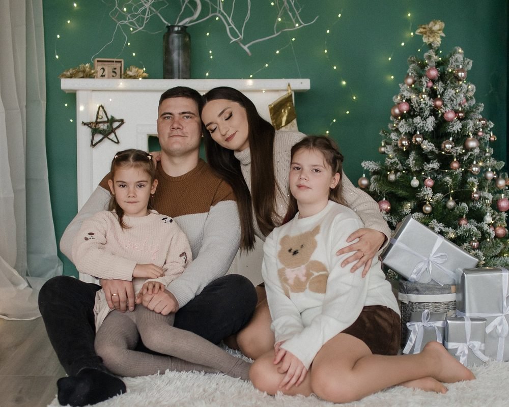 Šeima sėdi prie kalėdinės eglutės ir dovanų, fone žalia siena su girliandomis – kalėdinė fotosesija Šiauliuose