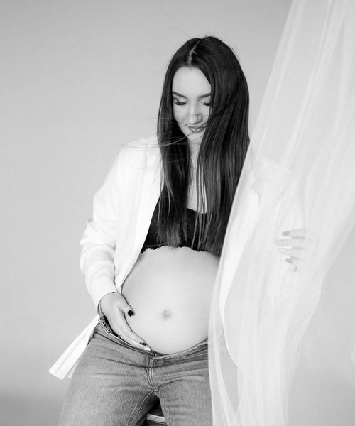 Nėščia moteris studijoje švelniai laiko pilvą, apsirengusi baltais marškiniais – laukimo fotosesija Šiauliuose