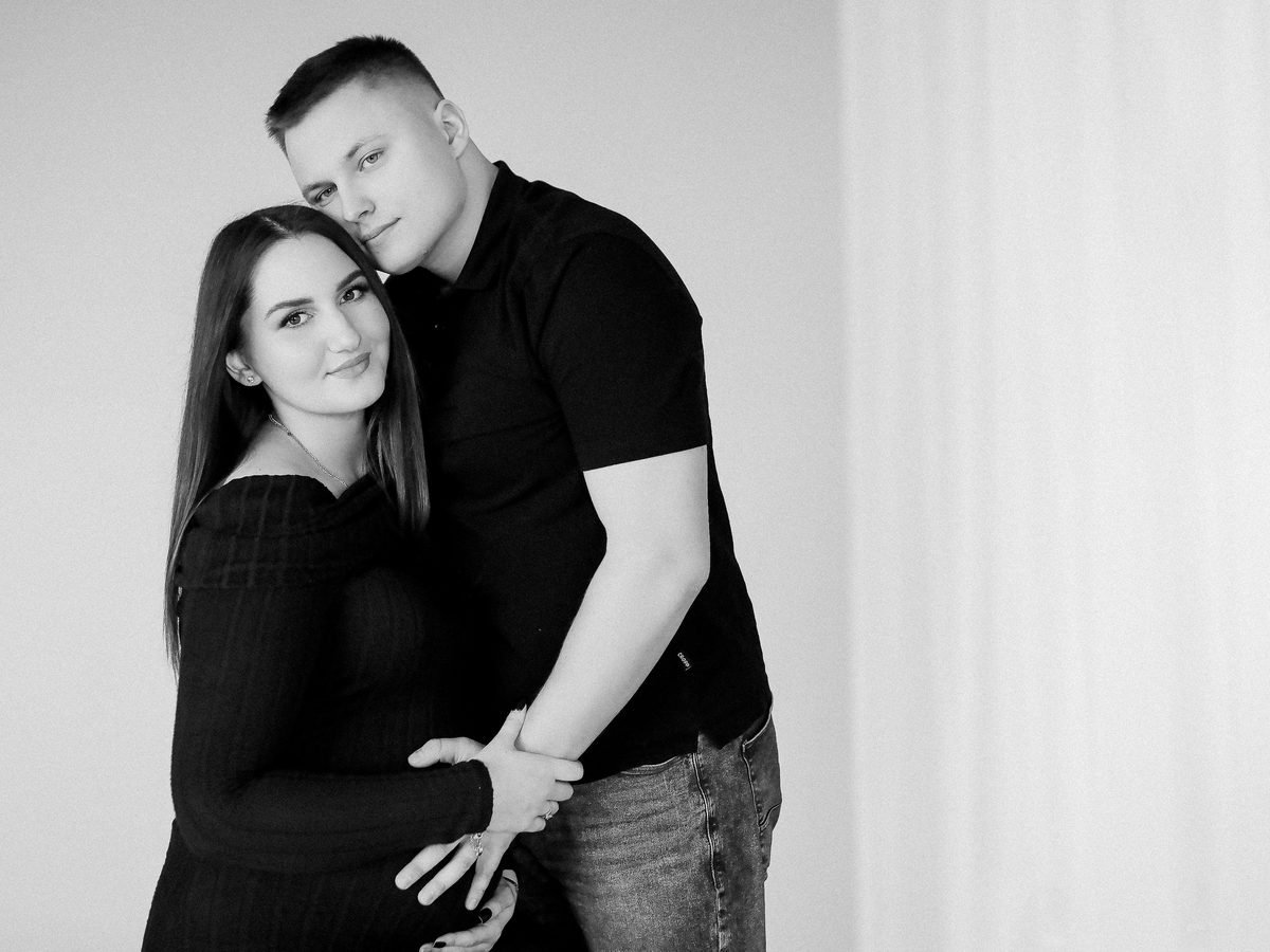 Nėščia moteris ir vyras apsikabinę studijoje, rankos ant nėštumo pilvuko – laukimo fotosesija Šiauliuose