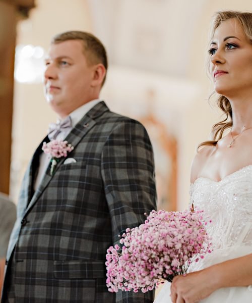 Jaunikis ir nuotaka stovi bažnyčioje su rausvų gėlių puokšte – vestuvių fotosesija Šiauliuose