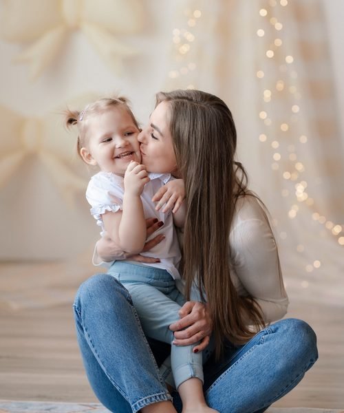 Mama bučiuoja mergaitę į skruostą sėdėdamos ant grindų – šeimos fotosesija Šiauliuose
