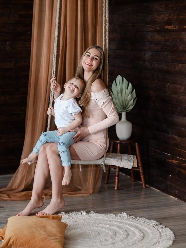 Mama su dukra sėdi ant sūpynių prie medinės sienos – šeimos fotosesija Šiauliuose