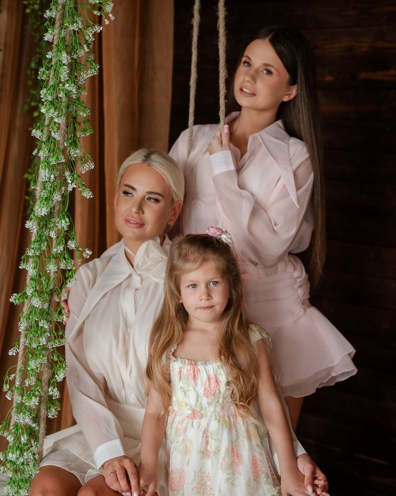 Dvi moterys ir mergaitė pozuoja prie sūpynių, papuoštų gėlėmis – šeimos fotosesija Šiauliuose