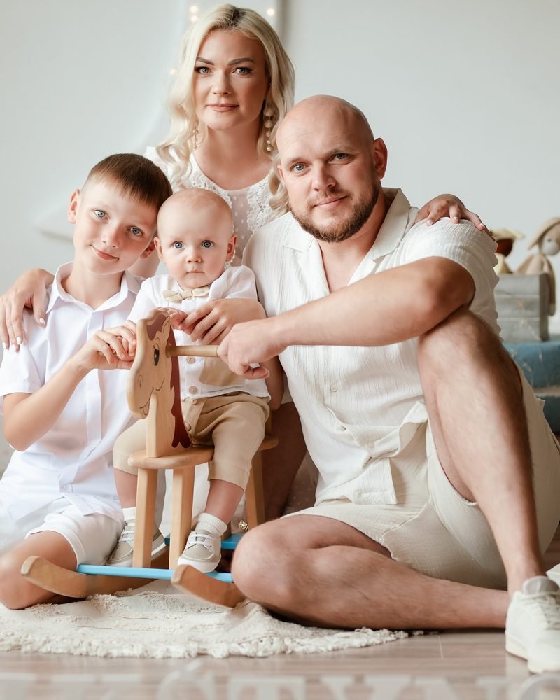 Šeima su dviem berniukais pozuoja studijoje šalia medinio arkliuko žaislo – krikštynų fotosesija Šiauliuose