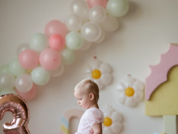 Mergaitė stovi tarp rausvų balionų ir gėlyčių sienos dekoro – cake smash fotosesija Šiauliuose