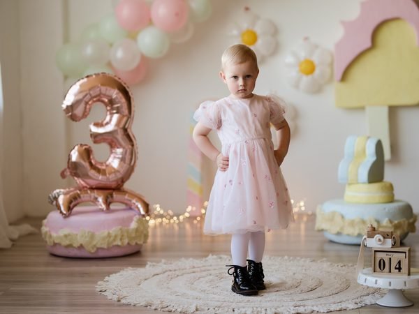 Mergaitė rausva suknele stovi tarp gimtadienio dekoracijų su balionu skaičiumi 3 – cake smash fotosesija Šiauliuose