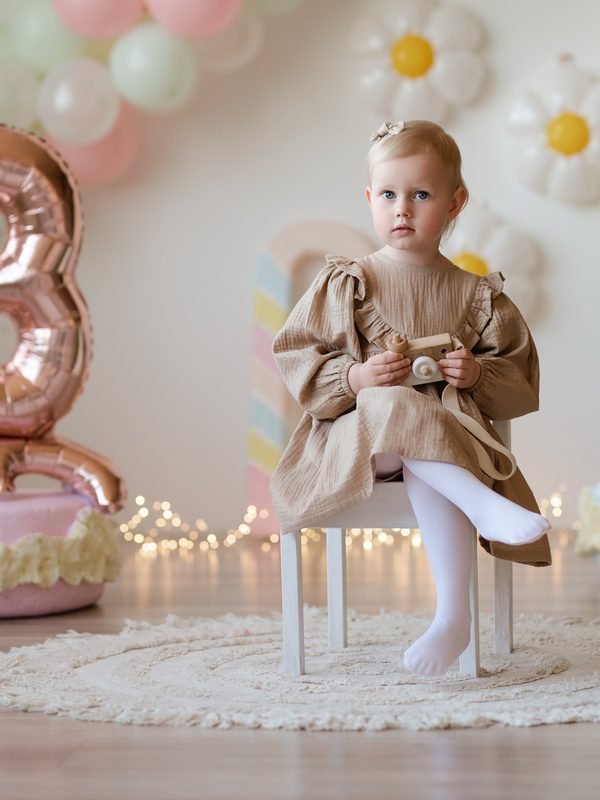 Mergaitė sėdi ant kėdutės su žaislyne kamera, aplink gimtadienio balionai – cake smash fotosesija Šiauliuose