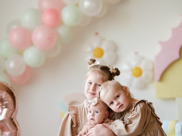 Trys mergaitės apsikabinusios pozuoja prie balionų arkos su gėlių dekoru – cake smash fotosesija Šiauliuose