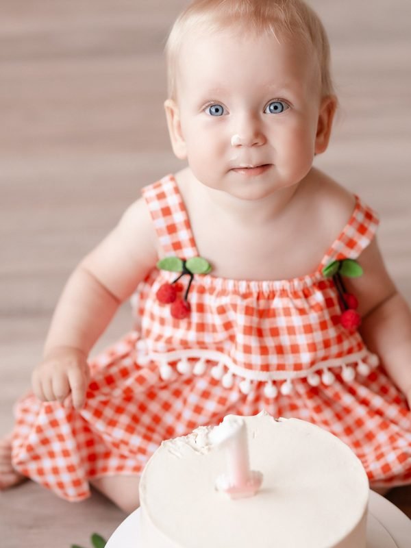 Kūdikis languota suknele su vyšniomis sėdi prie torto su žvakute – cake smash fotosesija Šiauliuose