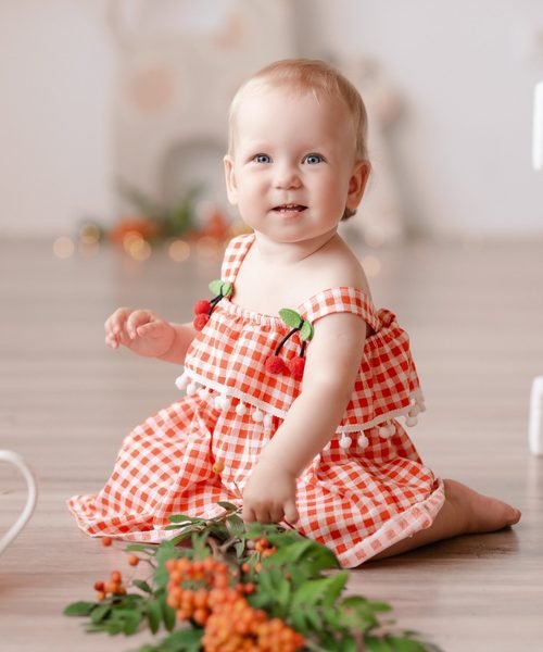 Kūdikė languota suknele sėdi ant grindų šalia raudonų uogų šakelių – cake smash fotosesija Šiauliuose