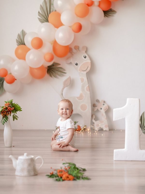 Kūdikis sėdi ant grindų tarp balionų ir žirafos dekoracijų – cake smash fotosesija Šiauliuose