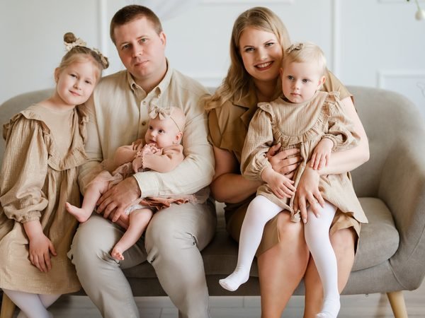 Vyresnioji dukra laiko kūdikį tėvų glėbyje sofoje vienodais rūbais – šeimos fotosesija Šiauliuose