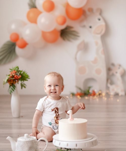 Kūdikis sėdi prie balto torto tarp balionų ir žirafos dekoro – cake smash fotosesija Šiauliuose