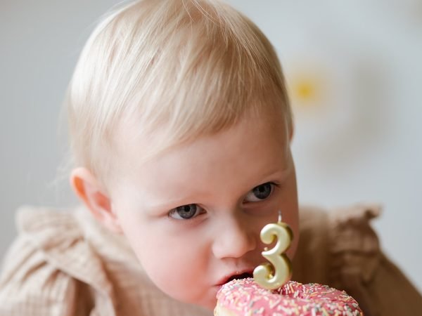 Vaikas valgo rausvą spurgą su žvake, vaizduojančia skaičių 3 – cake smash fotosesija Šiauliuose