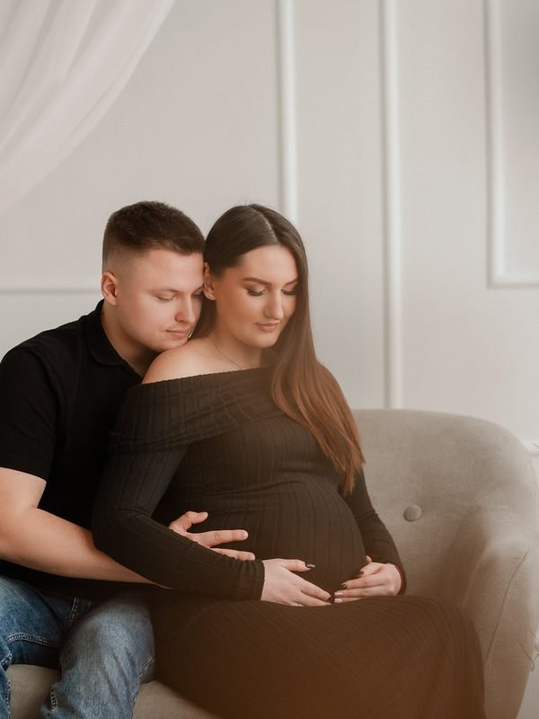 Vyras apkabina ant kelių sėdinčią nėščią moterį fotelyje – laukimo fotosesija Šiauliuose