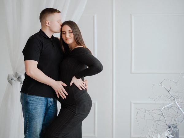 Vyras bučiuoja nėščią moterį į kaktą, rankos ant pilvo – laukimo fotosesija Šiauliuose
