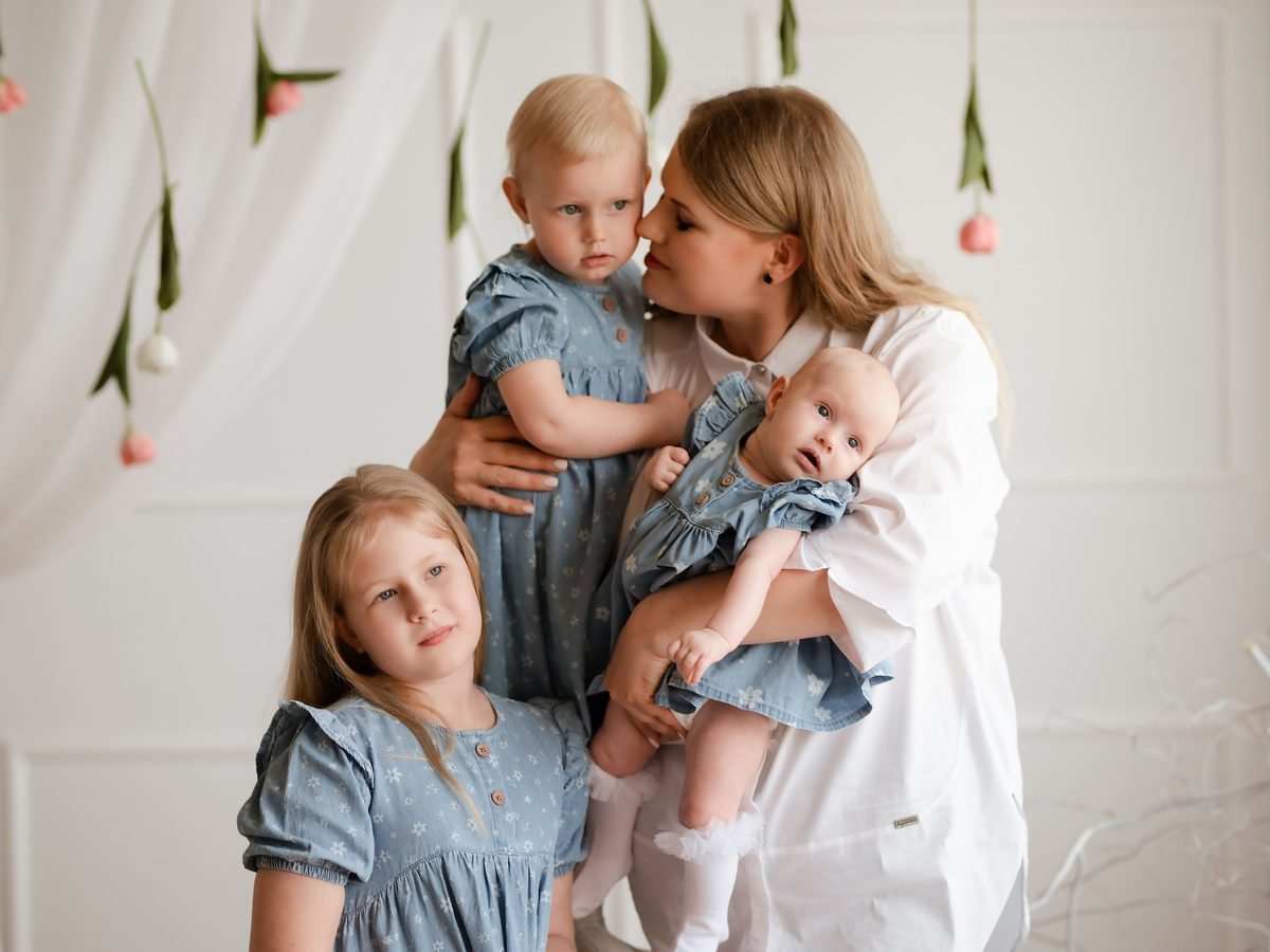 Mama su trimis dukromis pozuoja studijoje, kabo gėlių girliandos fonas – šeimos fotosesija Šiauliuose