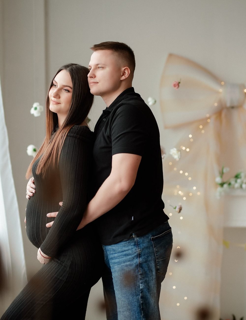 Nėščia moteris apsikabinusi su vyru stovi prie lango – laukimo fotosesija Šiauliuose