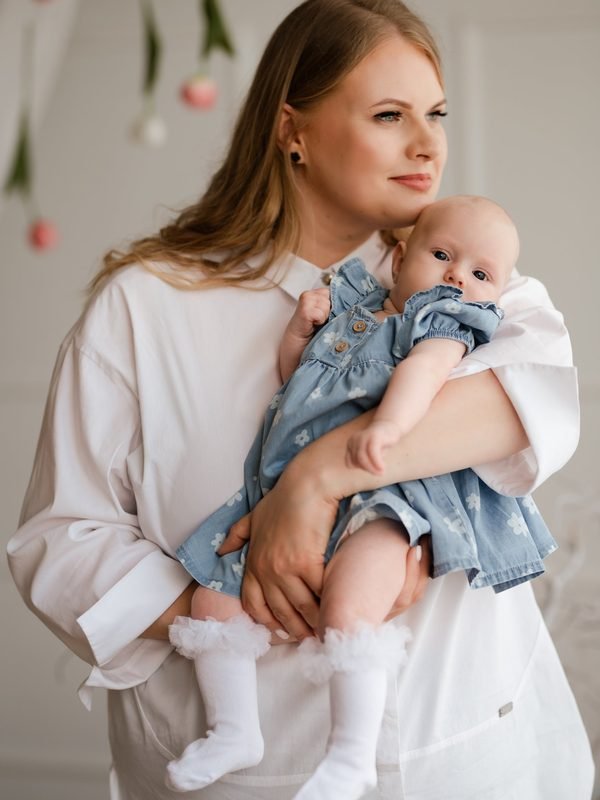 Mama laiko kūdikį ant rankų, žvelgia į šalį, kūdikis dėvi džinsinę suknelę – šeimos fotosesija Šiauliuose