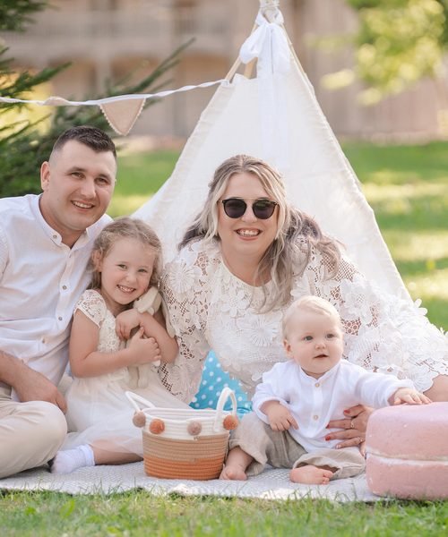 Šeima su dviem vaikais sėdi lauke prie balto tipi tento – krikštynų fotosesija Šiauliuose