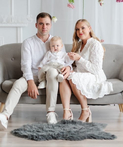 Tėvai su mažyliu sėdi ant sofos, apsirengę šviesiais drabužiais – krikštynų fotosesija Šiauliuose