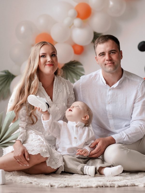 Šeima sėdi ant kilimo, kūdikis rankose laiko batuką, fone balionų arka – cake smash fotosesija Šiauliuose